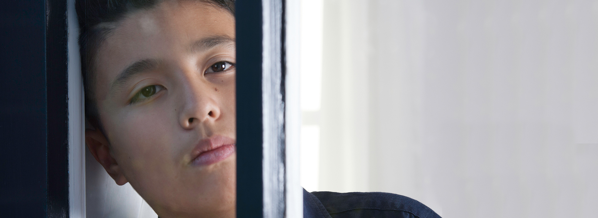 Boy leaning against window staring into distance thoughtful